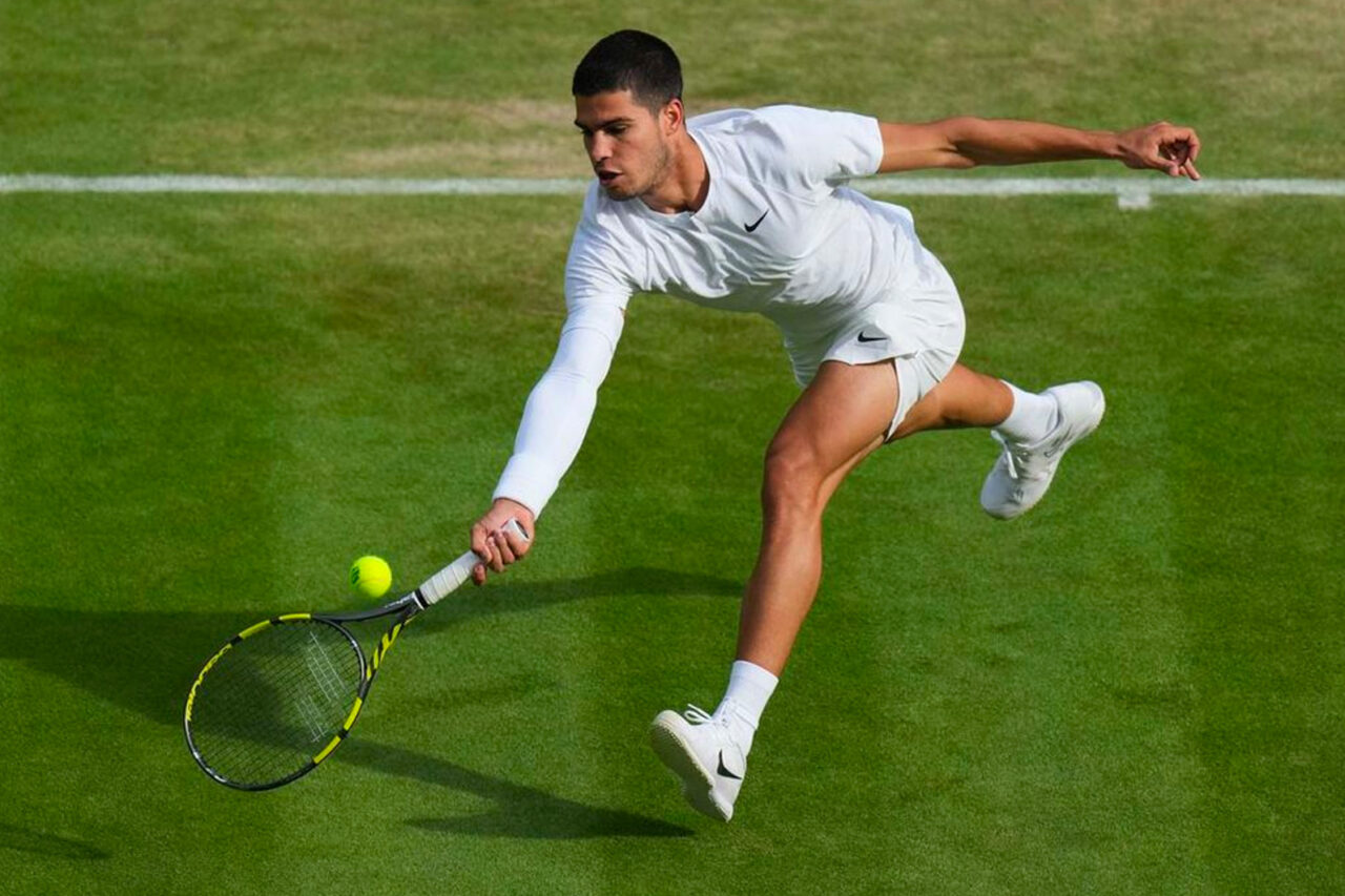 A male tennis player in white attire stretches to hit a low ball at the Wimbledon Championships, showcasing dynamic action on the iconic grass court.