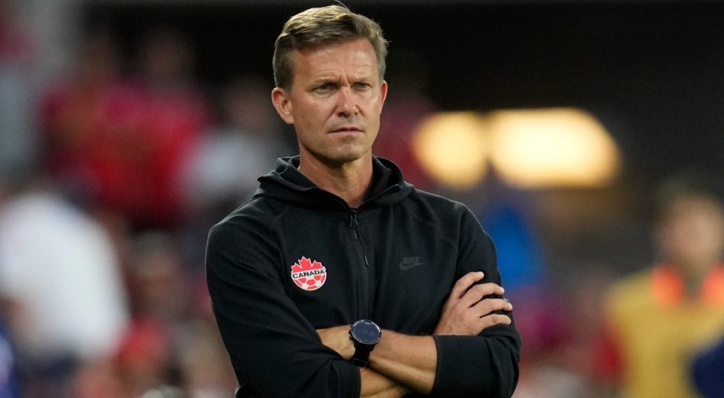 A Team Canada coach stands with his arms crossed, wearing a black Nike jacket with a Canada logo, intently watching a soccer game in a stadium.