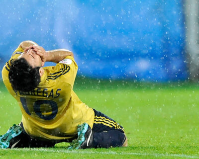 Soccer player in a yellow jersey sits on the field, holding his head in apparent despair or frustration, during a rainy match.