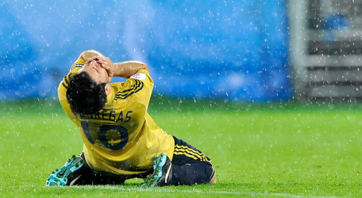 Soccer player in a yellow jersey sits on the field, holding his head in apparent despair or frustration, during a rainy match.