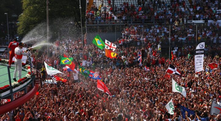 A Formula 1 driver sprays champagne at a cheering crowd with various national flags, capturing the vibrant celebration at the Italian Grand Prix.