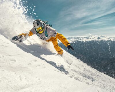 Snowboarder making a sharp turn on a snow-covered slope with a mountainous backdrop, related to Winter Games FAQ about hotels and tickets.