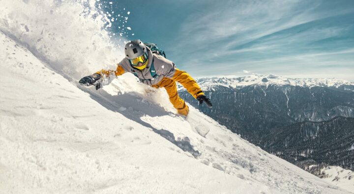 Snowboarder making a sharp turn on a snow-covered slope with a mountainous backdrop, related to Winter Games FAQ about hotels and tickets.