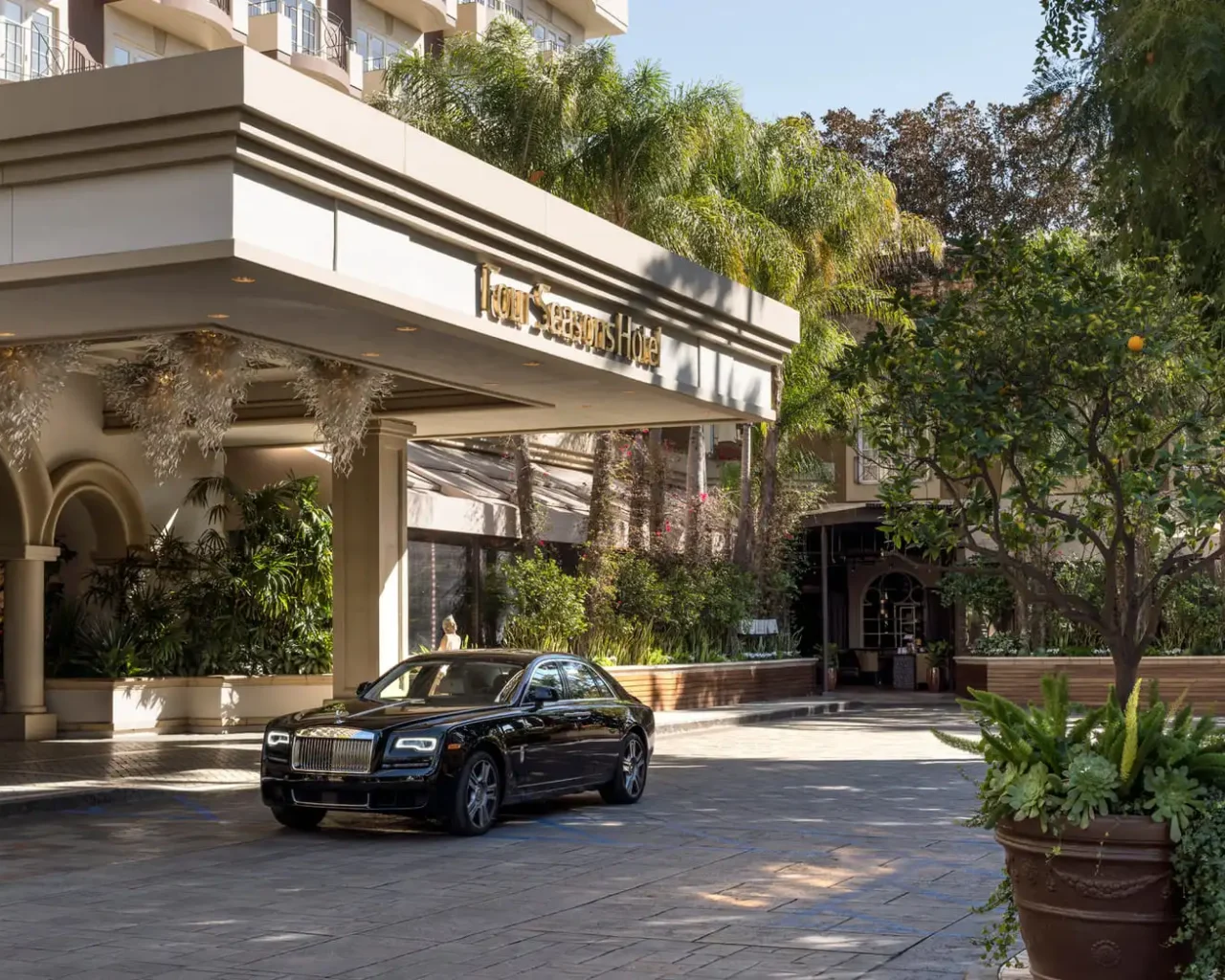 A luxury car parked under the portico of the Four Seasons Hotel in Beverly Hills, surrounded by lush greenery and sunny skies, ideal for Los Angeles Summer Games travel packages.