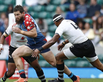 A rugby player in a star-spangled uniform runs with the ball, evading a tackle from an opponent wearing black and white stripes, ideal for those connecting through rugby travel packages.