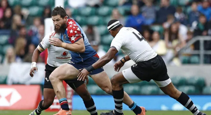 A rugby player in a star-spangled uniform runs with the ball, evading a tackle from an opponent wearing black and white stripes, ideal for those connecting through rugby travel packages.