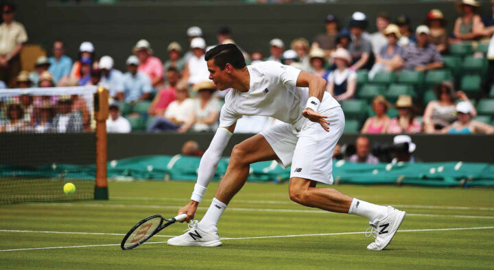A male tennis player in white attire stretches to hit a low-bouncing tennis ball on a grass court, exemplifying the dynamic action often seen in tennis travel packages.