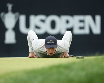Golfer lies flat on his stomach, lining up a putt on the green, with the US Open logo prominently displayed in the background, capturing a moment at the prestigious golf tournament.