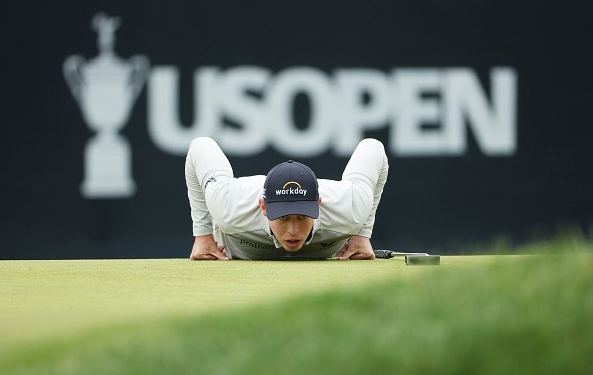 Golfer lies flat on his stomach, lining up a putt on the green, with the US Open logo prominently displayed in the background, capturing a moment at the prestigious golf tournament.