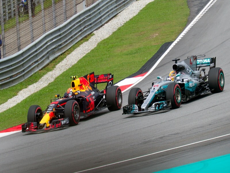 Two Formula 1 cars race closely on a track, creating an intense competitive atmosphere at the Spanish Grand Prix; one car is decorated in vibrant red and yellow, the other in silver and teal.