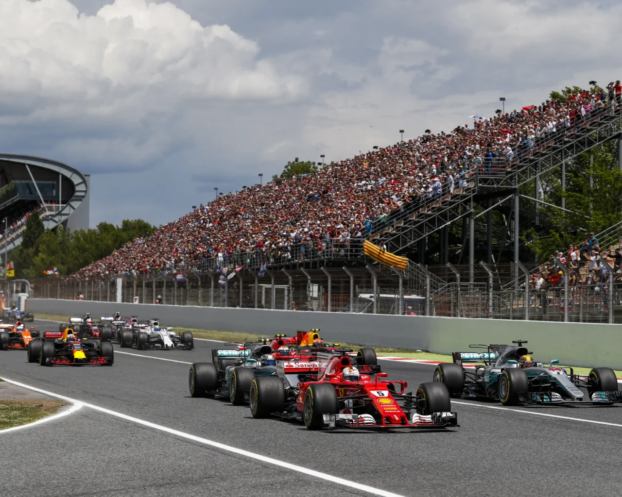 Formula 1 cars race on the track with a packed audience in the grandstands at the Spanish Grand Prix, promoting the Roadtrips travel package.