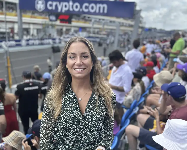 A smiling woman stands at the Miami Grand Prix, with spectators and the race track in the background, highlighting the vibrant atmosphere of the event.