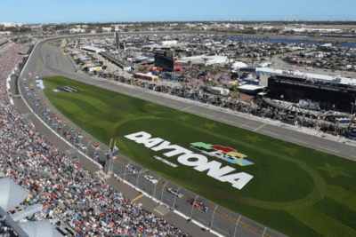 Aerial view of Daytona International Speedway showing a crowded grandstand and race track during a major event, with a large Daytona logo visible on the infield grass.