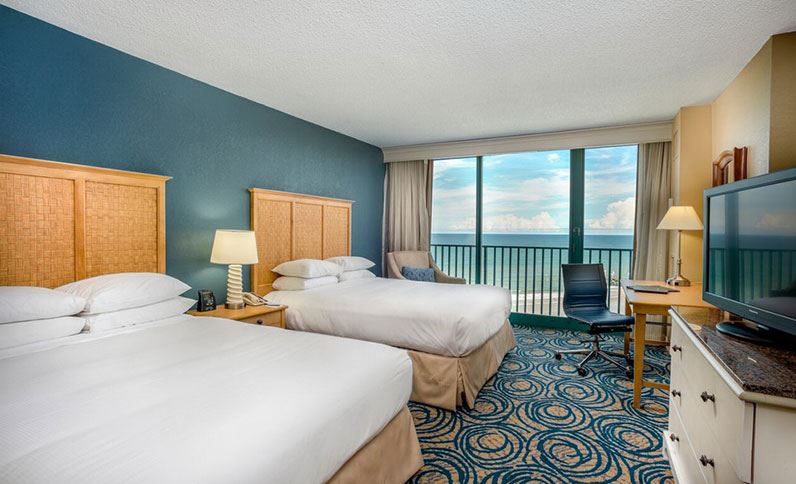 A well-appointed hotel room at Hilton Daytona Beach Oceanfront Resort, featuring two beds, a desk, and a large window overlooking the ocean, optimal for Daytona 500 accommodations.