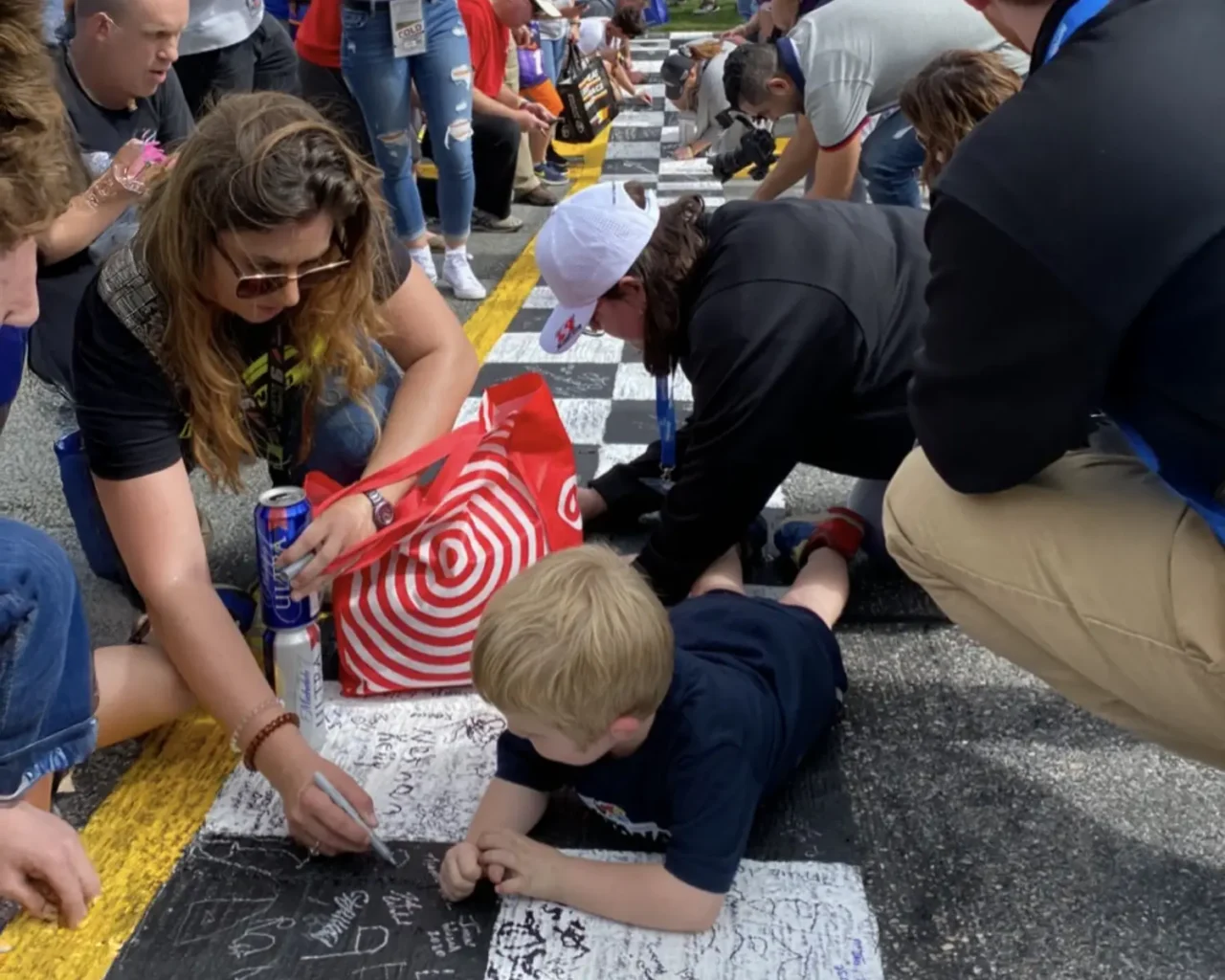 Fans, including children and adults, are writing and drawing with chalk on the race track at the Daytona 500 Fanzone, enhancing their event experience with preferred tickets and pre-race access.