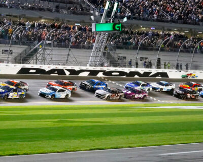 Racing cars speed around the Daytona track during an evening race, with a crowd of spectators watching intently from the stands.