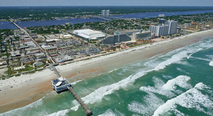 Aerial view of Daytona Beach, showcasing its expansive shoreline, bustling streets, and a pier extending into the ocean, epitomizing a typical sunny day perfect for discovering road trips and local events.