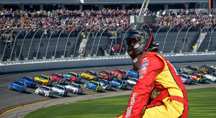 A racing driver in a red suit watches as a group of colorful NASCAR cars compete at Daytona International Speedway in Florida, surrounded by a large crowd of enthusiastic fans.