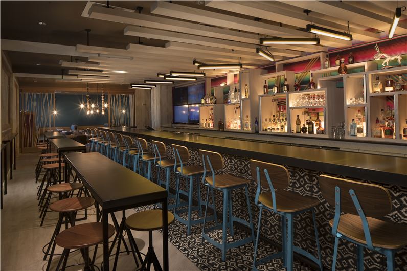 Modern bar area at the Hilton Daytona Beach Oceanfront Resort featuring stylish stools, a long counter, and a vibrant backlit display of bottles, ideally located near Daytona 500 event accommodations.
