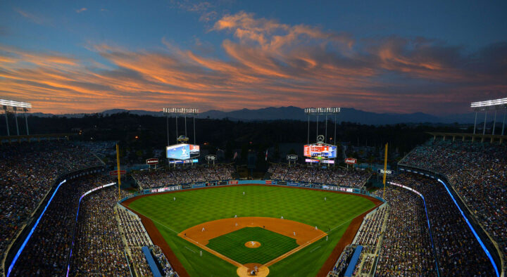 View of a baseball stadium packed with spectators during a game at dusk, with a colorful sunset sky in the background, ideal for MLB All-Star travel package promotions.