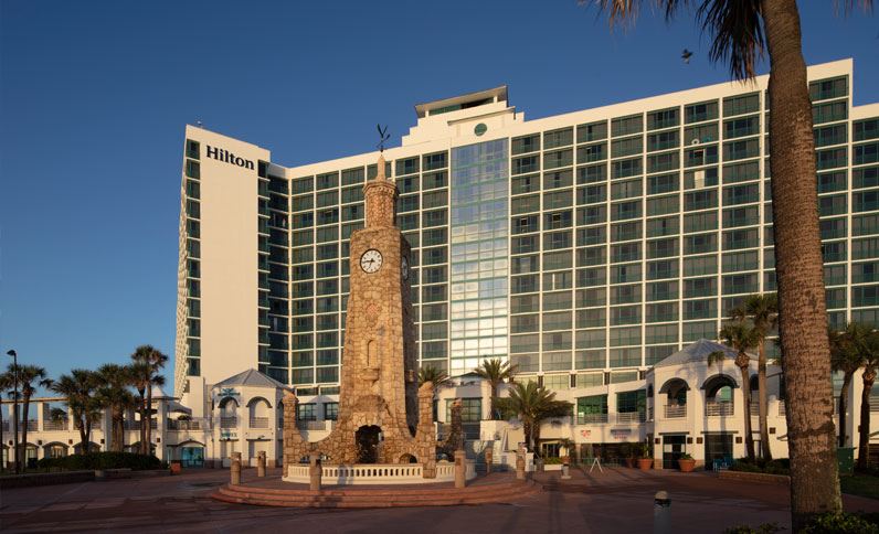 Hilton Daytona Beach Oceanfront Resort, a luxury hotel in Daytona, Florida, prominently features a clock tower in a sunny, palm-lined setting near the beach.