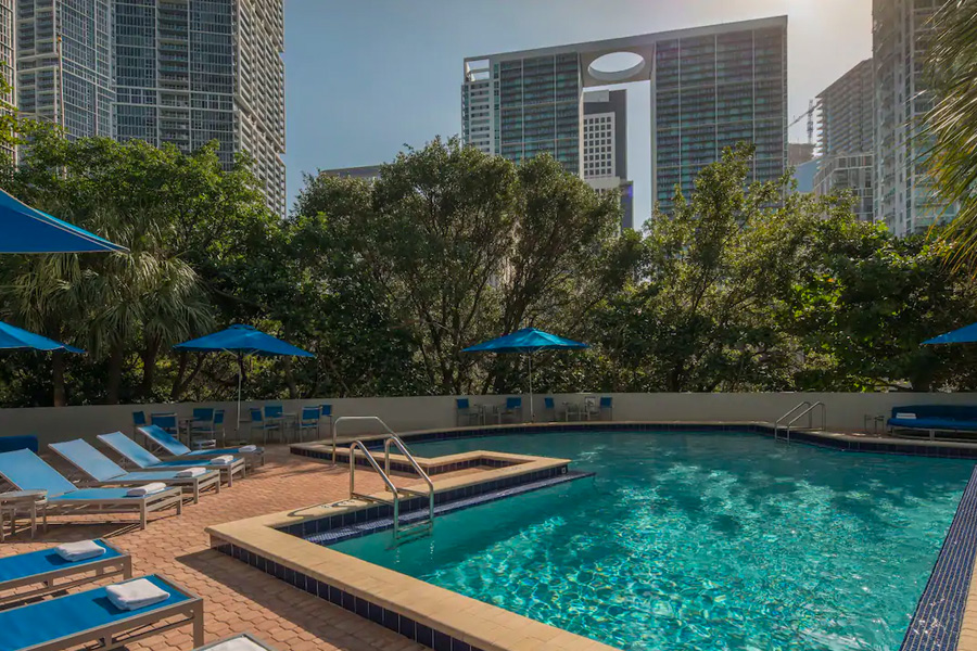 Outdoor pool area at Hyatt Regency Miami surrounded by blue lounge chairs and shaded by blue umbrellas, nestled among high-rise buildings, ideal for Miami Grand Prix hotels & accommodations.