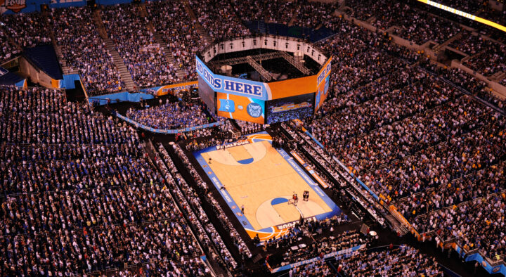 An aerial view of a crowded Lucas Oil Stadium during a Final Four basketball game, featuring prominently displayed banners and a uniquely designed court.