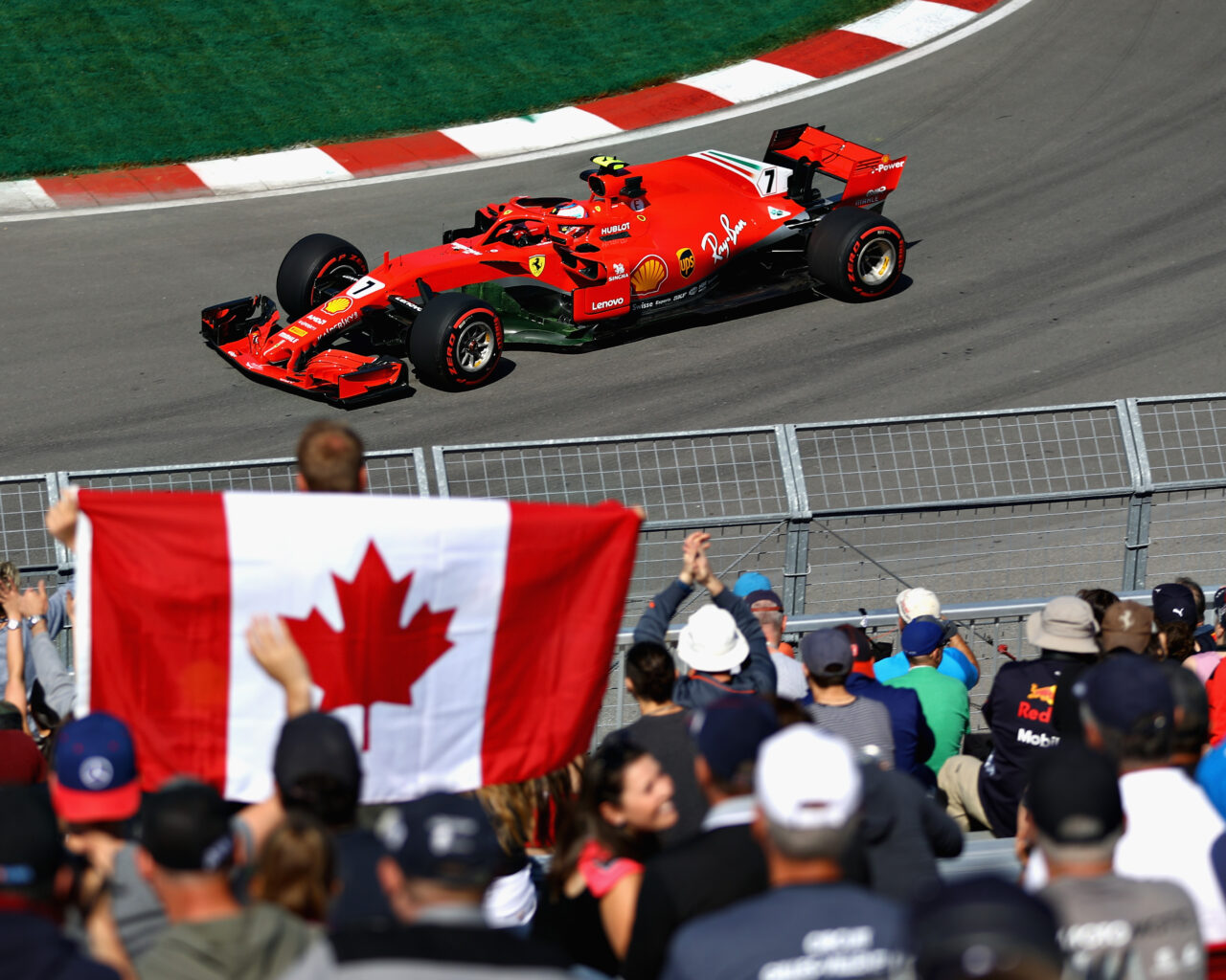 A red racing car speeds along the track at the Canadian Grand Prix, watched by a crowd waving a large Canadian flag.