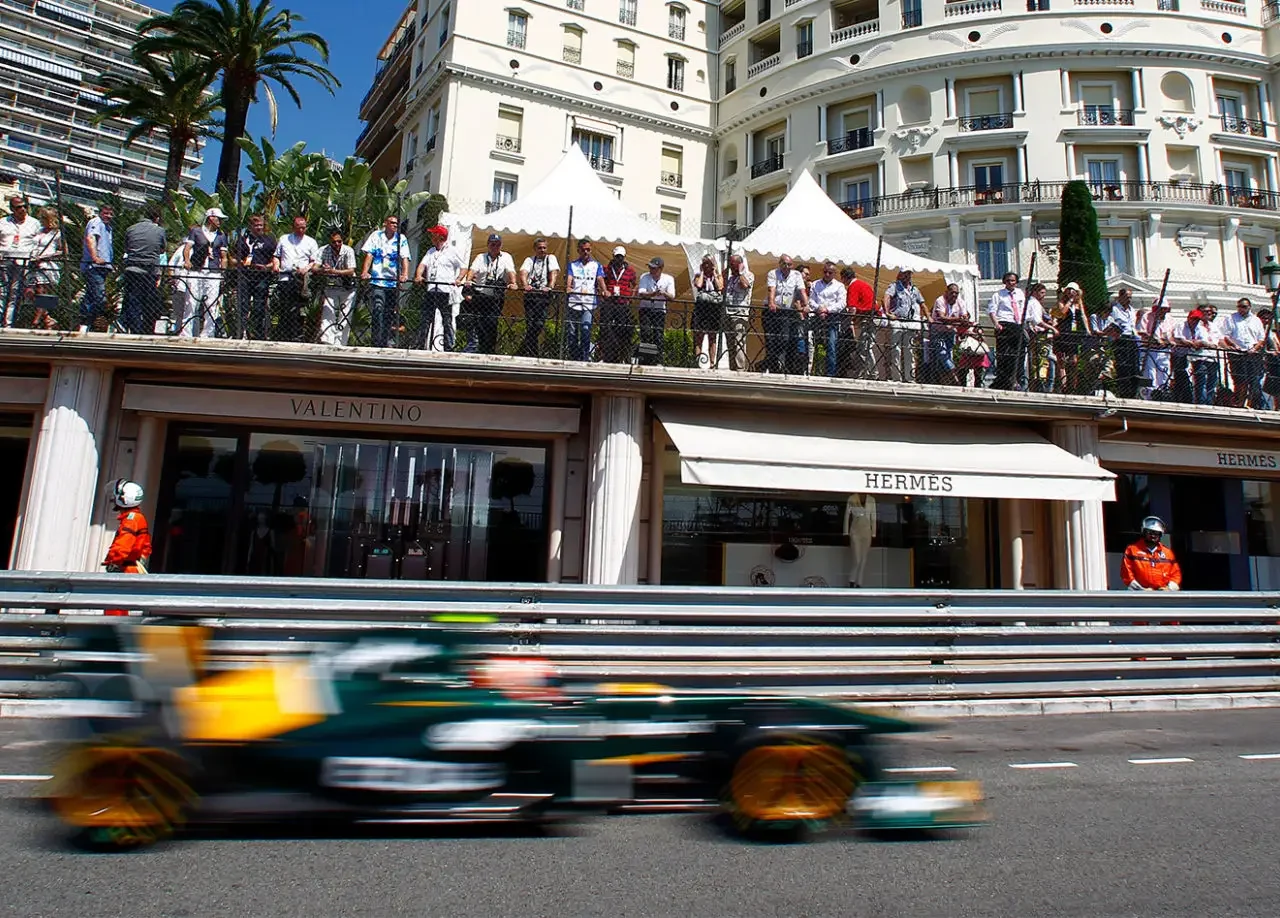A high-speed Formula One car zooms past spectators on a Monaco city street, lined with luxury boutiques like Valentino and Hermès, offering an immersive view of the Monaco Grand Prix event.