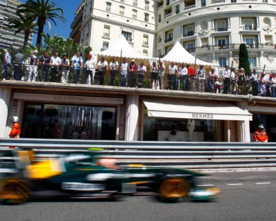A high-speed Formula One car zooms past spectators on a Monaco city street, lined with luxury boutiques like Valentino and Hermès, offering an immersive view of the Monaco Grand Prix event.