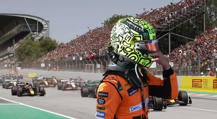 Racing driver in a vibrant helmet watches competitors at Circuit de Barcelona-Catalunya, a notable setting for Grand Prix auto racing travel packages.