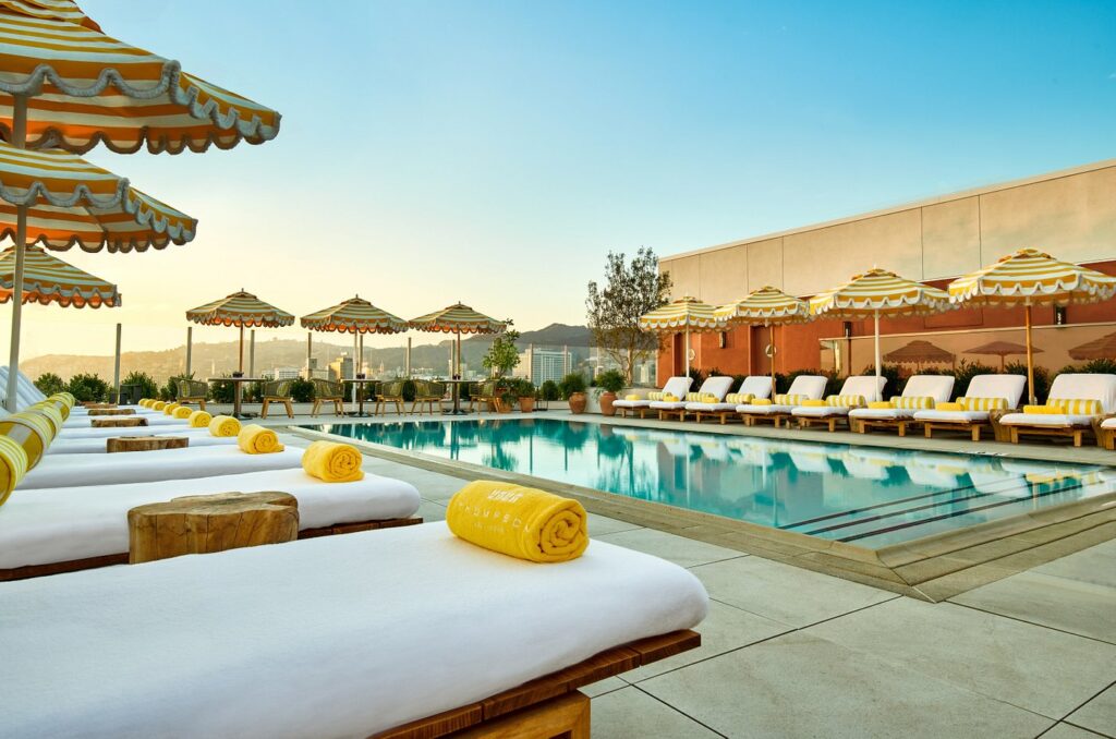 Luxurious outdoor pool area at Thompson Hollywood by Hyatt, featuring lined-up sun loungers and striped umbrellas, catering well to tourists visiting Los Angeles for Summer Games events.