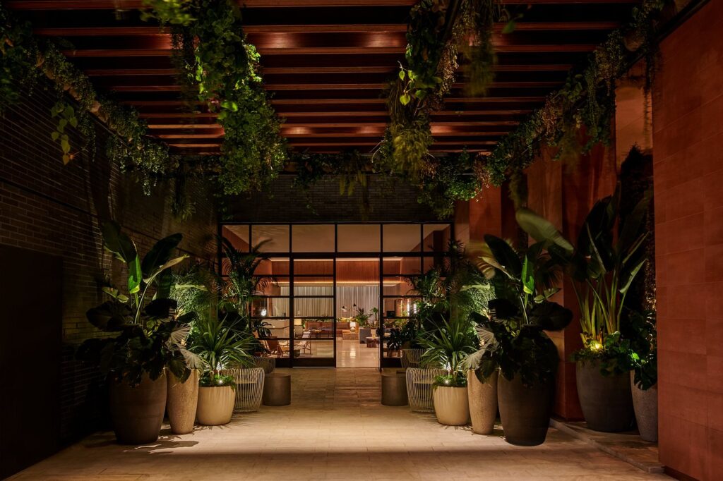 Nighttime view of the Thompson Hollywood hotel's lush, plant-lined walkway, illuminated with warm lighting, leading to a modern glass-fronted entrance, ideal for Summer Games accommodations in Los Angeles.