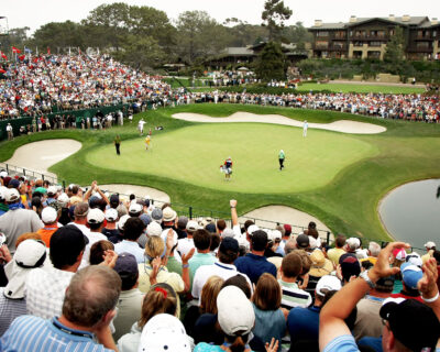 A golf player completes a putt as a large crowd watches at the 18th hole by a scenic pond, highlighting an iconic moment at a US Open Golf event, ideal for custom travel packages.