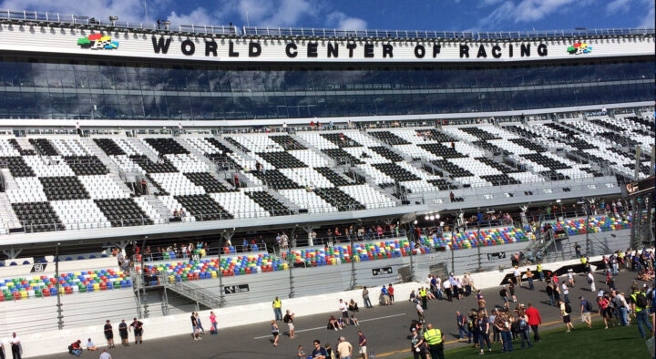 View of the Daytona International Speedway, showing crowded stands and spectators on the track, with the 'WORLD CENTER OF RACING' signage displayed prominently above.