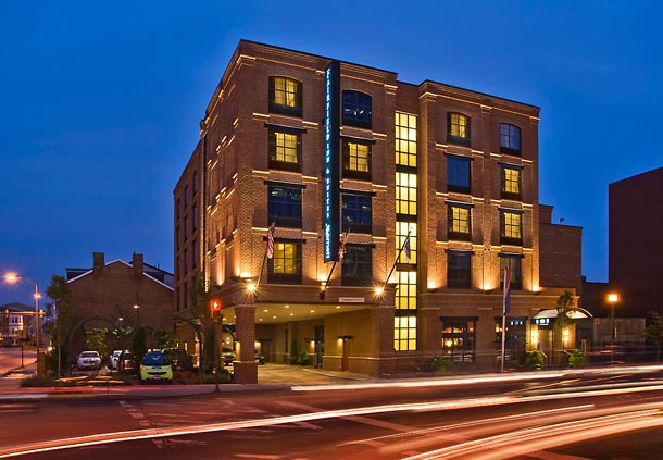 Fairfield Inn & Suites Baltimore Downtown/Inner Harbor at twilight, with the building's lights on and blurred vehicle lights moving past, is near accommodations for the Preakness Stakes.