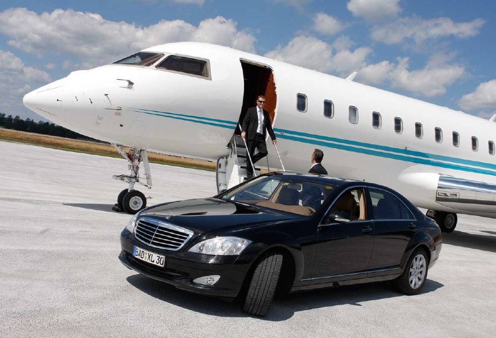 A man exits a private jet and approaches a luxury sedan on a sunny airport tarmac, symbolizing high-quality travel experiences and personalized pre-trip assistance.