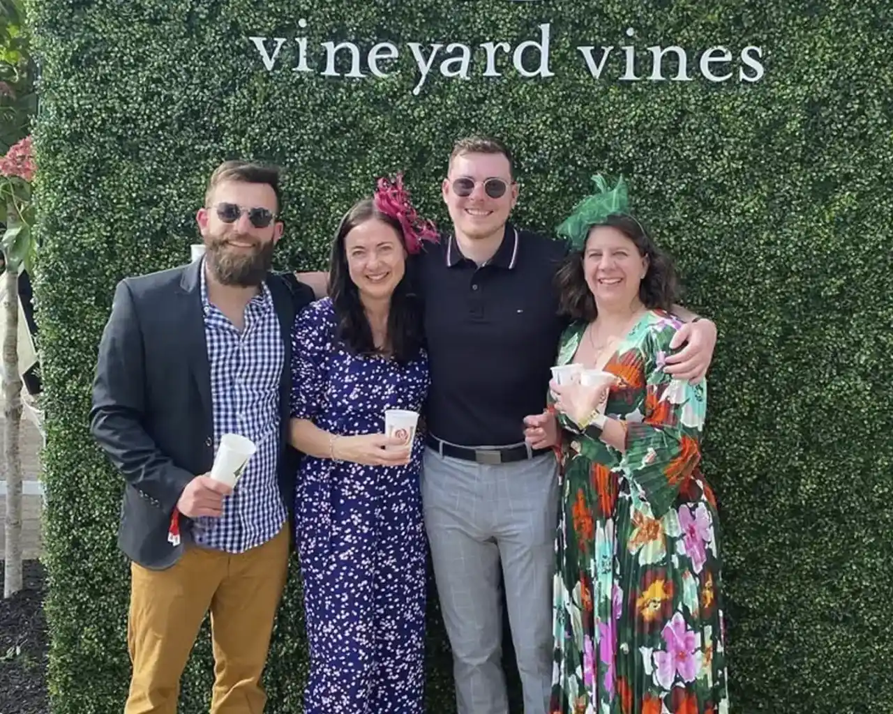 Two men and two women pose for a photo, smiling and dressed festively, in front of a 'vineyard vines' hedge at a Kentucky Derby-related event, highlighting premium tickets and travel packages.