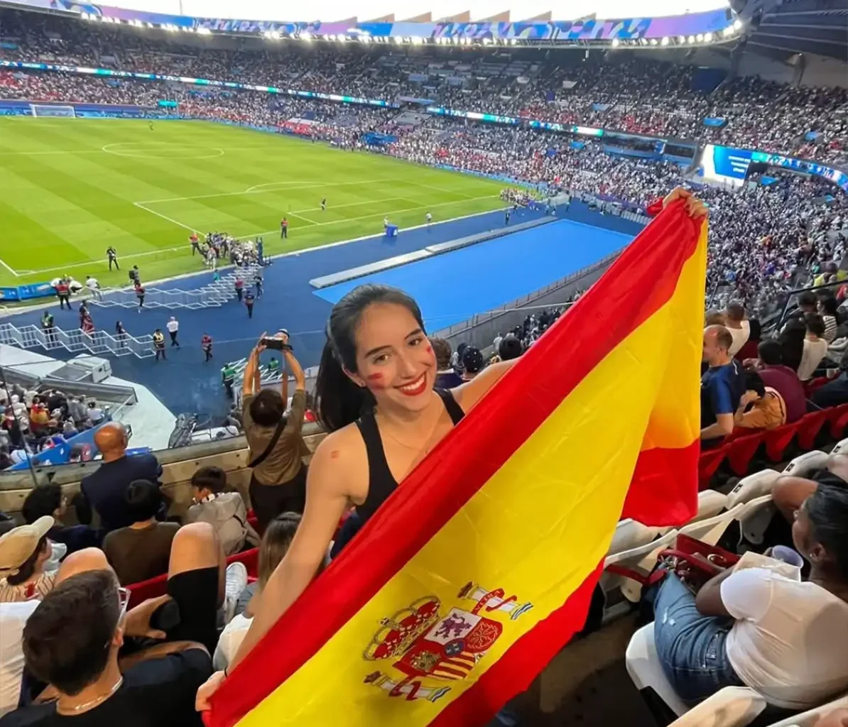 A woman holding a Spanish flag smiles at the camera in a crowded stadium during a Champions League match, enhancing the festive atmosphere of the event.