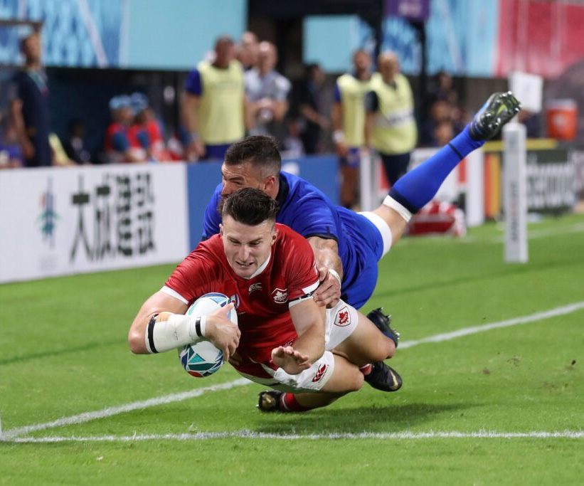 Two rugby players in a dynamic tackle during a match, with the player in red scoring a try as part of a custom World Cup Rugby travel package experience.