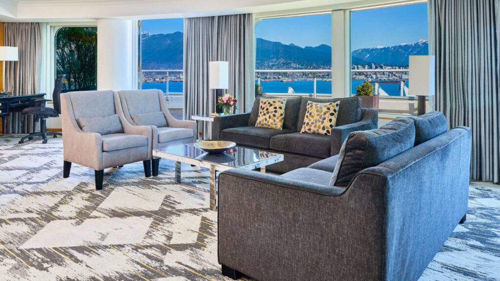 Luxurious hotel suite at the Pan Pacific Vancouver showing a stylish living area with modern furniture and a panoramic view of mountains and the waterfront, ideal for World Cup travel packages.