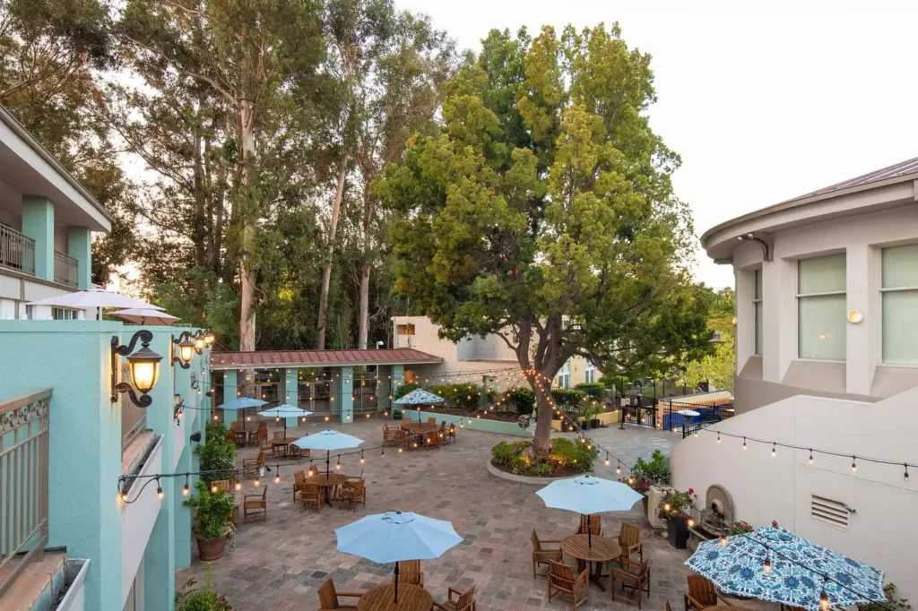 Outdoor dining area at Cabana Hotel in Palo Alto, showing tables with blue umbrellas surrounded by trees, ideal for visitors looking for luxury hotel accommodations near San Jose.
