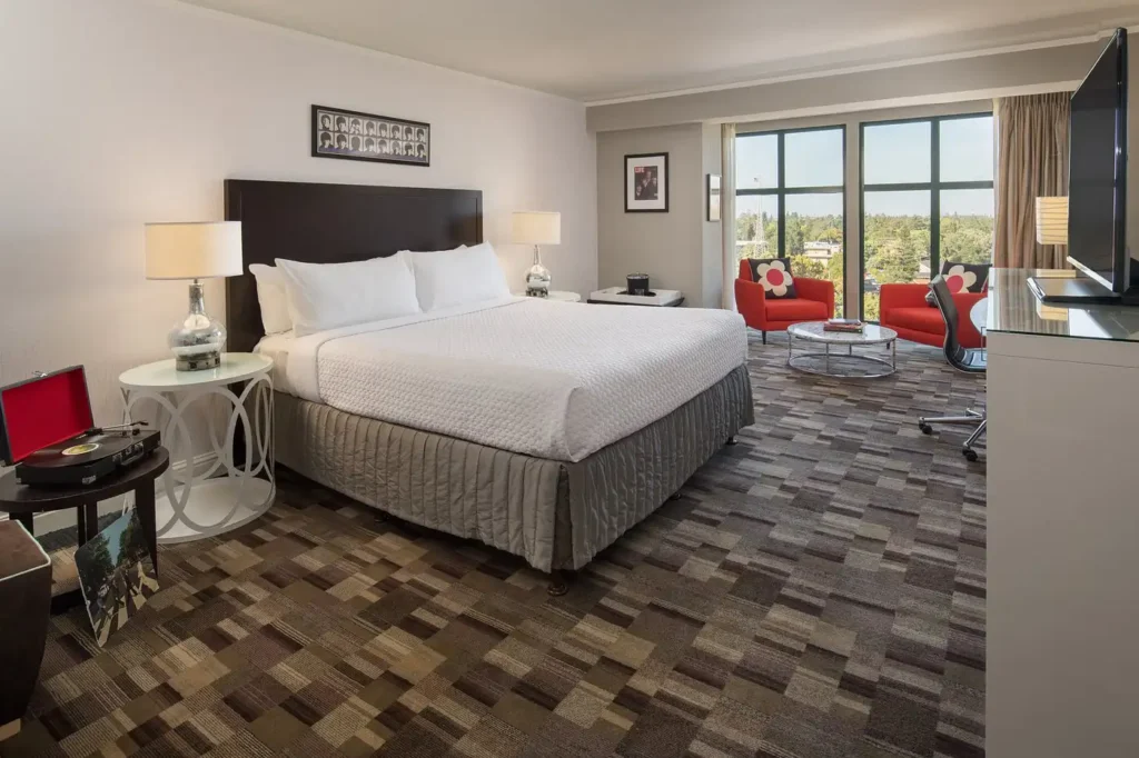 Interior view of a spacious room at the Cabana Hotel Palo Alto, featuring a large bed, seating area with bright red chairs, and expansive windows overlooking the surroundings, ideal for Palo Alto visitors.