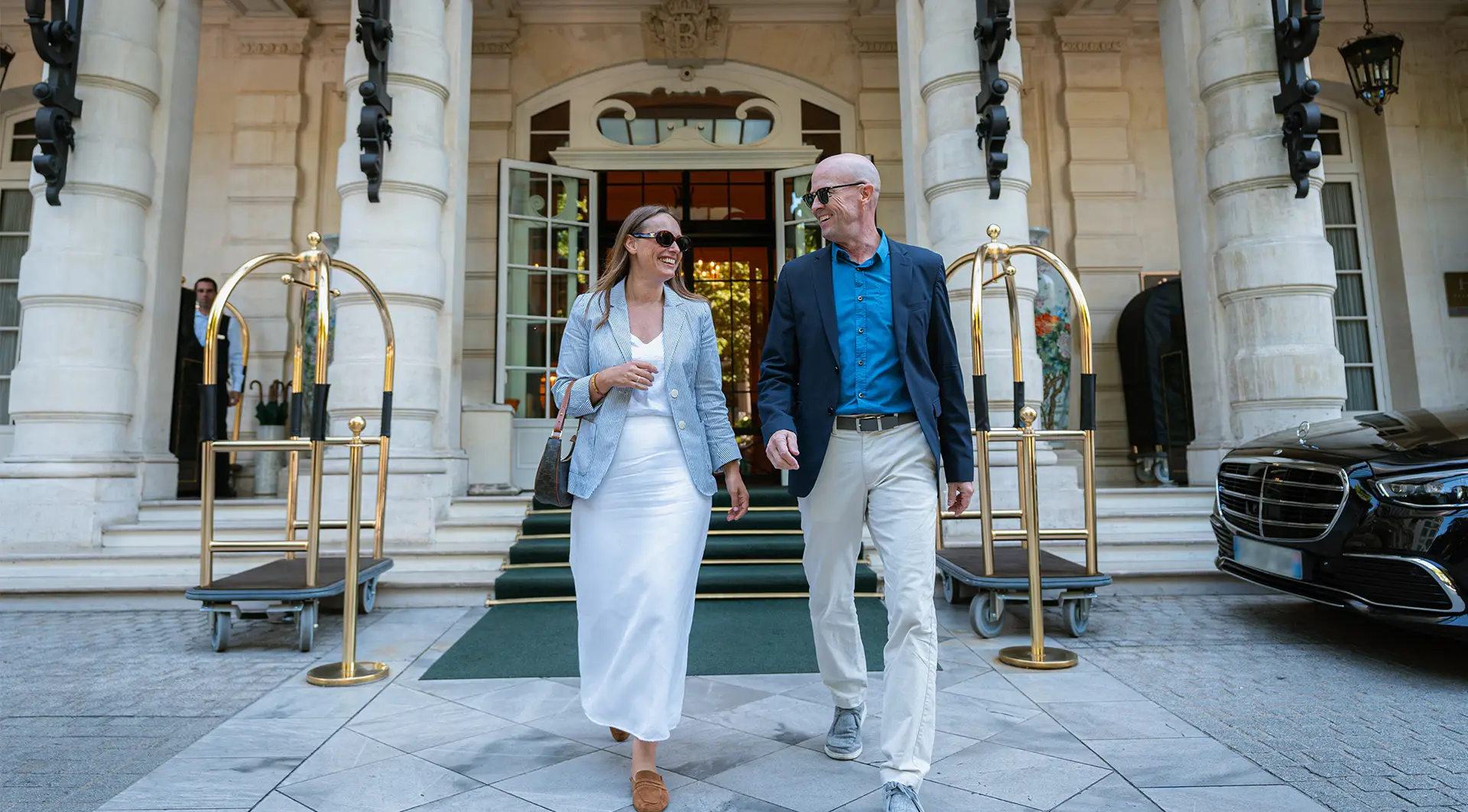 A mature couple happily exits a luxury hotel, engaging in conversation, representing a high-end sports travel package experience.