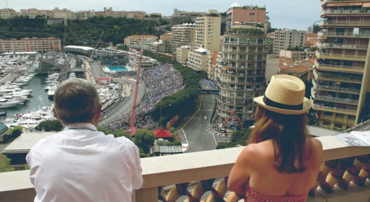 Two people view a bustling race event from a terrace in Monaco, showcasing a scene of crowded streets and docked yachts, ideal for groups seeking exclusive hospitality experiences.