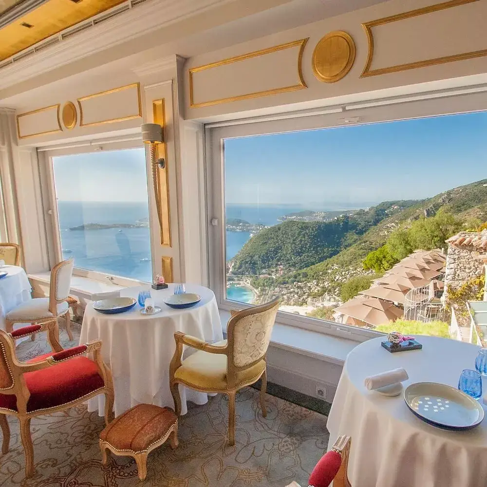 Elegant restaurant interior with tables set for dining, showcasing a panoramic view of the sea and surrounding hills through large windows, offering a high-quality dining experience.
