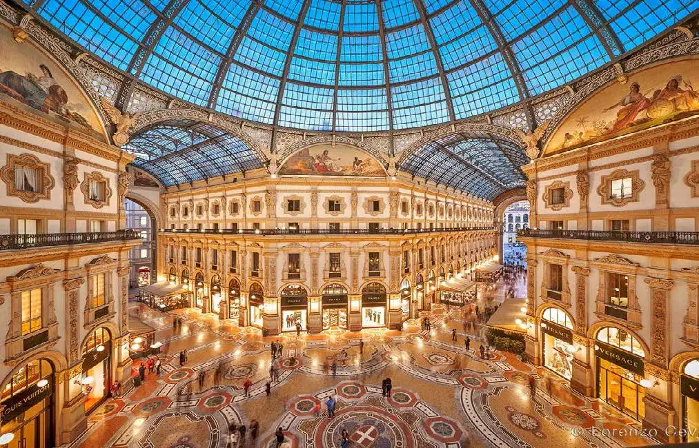 Inside the ornate Galleria Vittorio Emanuele II in Milan, shoppers stroll under a glass dome, surrounded by luxury shops and intricate mosaics, highlighting Milan's architectural and cultural richness.