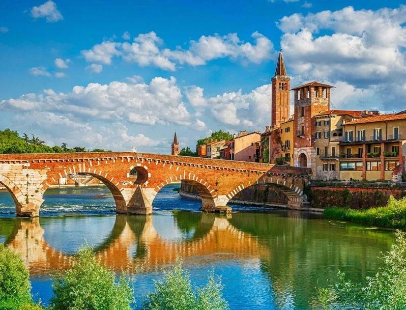 Ponte Pietra, the stone bridge in Verona, reflects in the river, surrounded by historical buildings under a sunny sky, highlighting the city's appeal for the Winter Games add-ons and Milan tours.